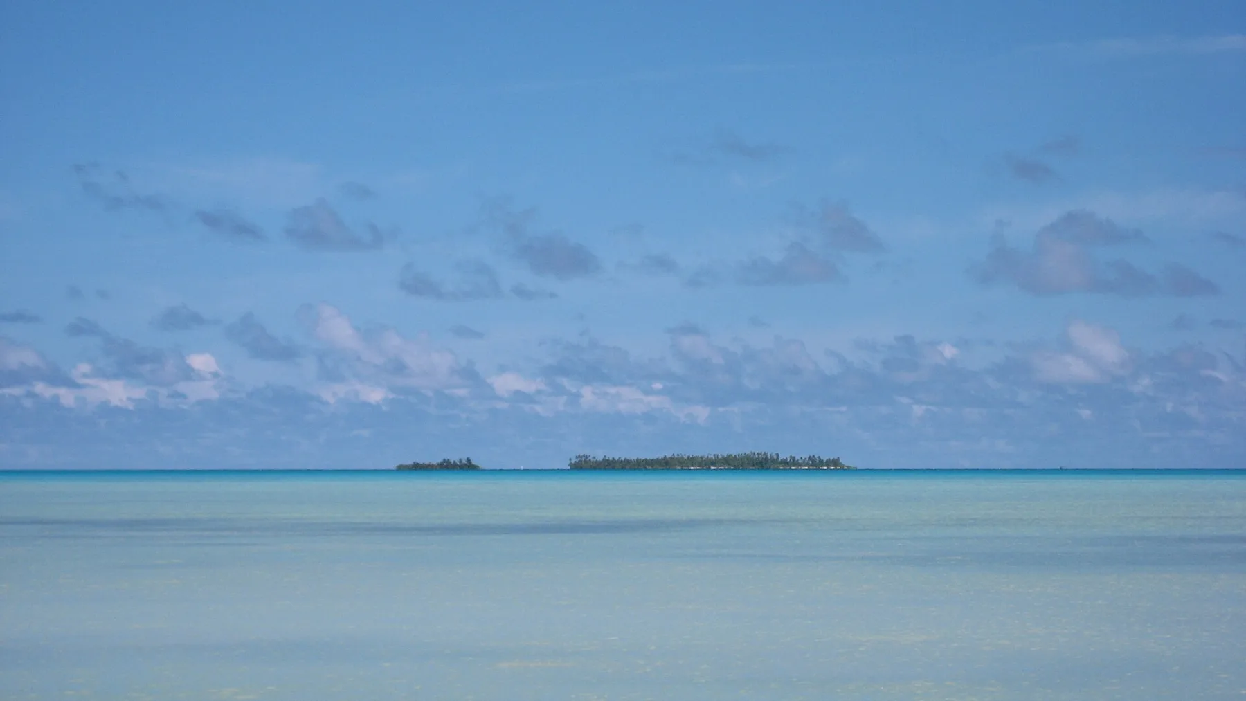 Twee kleine motu drijven als groene stipjes boven het spiegelgladde turquoise water van de Aitutaki-lagune