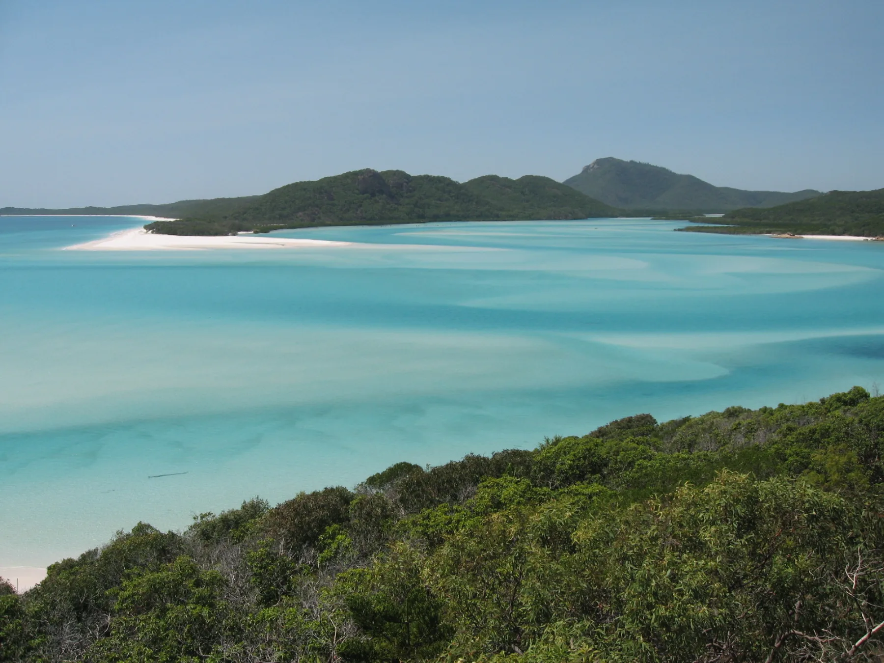 Vogelperspectief over Hill Inlet: witte silica-zandtongen wervelen door turquoise water, met de groene heuvels van Whitsunday Island op de achtergrond